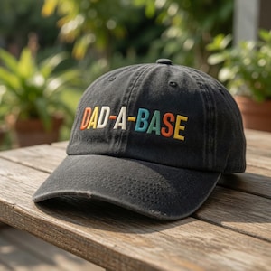 May include: A black baseball cap with the embroidered text "DAD-A-BASE" in colorful letters. The cap has a distressed look and a curved brim. The hat is sitting on a wooden surface.