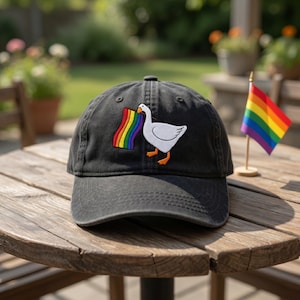 May include: A black baseball cap with a white goose design holding a rainbow flag. The cap is sitting on a wooden table, with a small rainbow flag in the background.