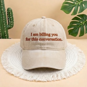 May include: A beige baseball cap with the embroidered text, "I am billing you for this conversation." The hat is sitting on a white, fringed surface, with a cactus and green leaves in the background.