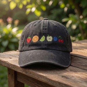 May include: A black baseball cap with embroidered fruit designs, including a strawberry, orange slice, lime wedge, apple, blueberry, and cherries. The cap is made of a soft, worn fabric and sits on a wooden surface.