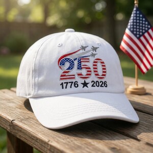 May include: White baseball cap with embroidered design. The design includes the number "250" in red and blue, the years "1776" and "2026", and a star. Above the number are three fighter jets. An American flag is in the background.