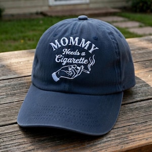 May include: Navy blue baseball cap with the embroidered text "MOMMY Needs a Cigarette" in white. The design includes a hand holding a lit cigarette. The cap has a curved brim and a fabric strap closure.