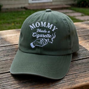 May include: Olive green baseball cap with the embroidered text "MOMMY Needs a Cigarette" in white. The design includes a hand holding a cigarette with smoke rising. The cap has a curved brim and a top button.