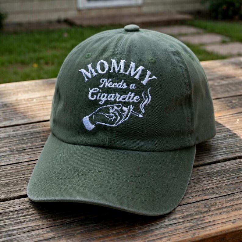May include: Olive green baseball cap with the embroidered text "MOMMY Needs a Cigarette" in white. The design includes a hand holding a cigarette with smoke rising. The cap has a curved brim and a top button.
