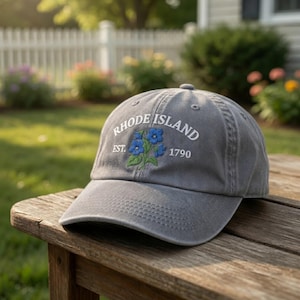 May include: Gray baseball cap with the text "RHODE ISLAND EST. 1790" embroidered in white. The cap features a floral design with blue flowers and green leaves. The hat is sitting on a wooden surface.