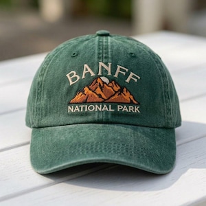 May include: A forest green baseball cap featuring an embroidered design of brown mountains with a white sun. The text BANFF is arched above the mountains, and the words NATIONAL PARK are embroidered in white thread below the mountain graphic.