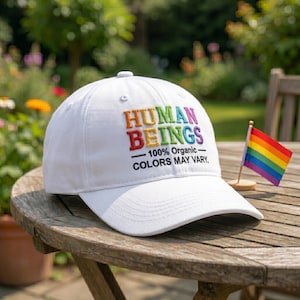 May include: White baseball cap with the embroidered text "HUMAN BEINGS" in rainbow colors. The cap also features the text "100% Organic - COLORS MAY VARY." A small rainbow flag is next to the cap.