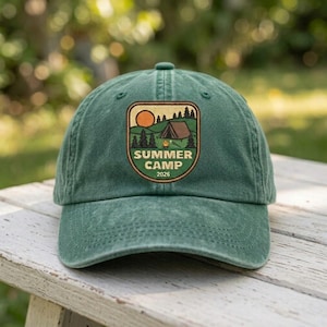 May include: A forest green baseball cap featuring an embroidered patch on the front. The patch displays a landscape with a brown tent, pine trees, and an orange sun. The text reads SUMMER CAMP 2026 in a bold, sans-serif font on the patch.
