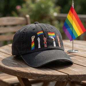 May include: A black baseball cap with embroidered rainbow flag and hand gestures. The cap features a curved brim and is sitting on a wooden table. A small rainbow flag on a stand is also visible.
