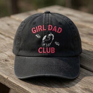 May include: Black baseball cap with the words "GIRL DAD CLUB" embroidered in pink. A white unicorn graphic is centered below the text. The cap is made of a soft, worn material.