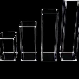 Könnte beinhalten: Vier transparente Acryl-Präsentationsständer in verschiedenen Höhen. Jeder Ständer hat die Form einer transparenten, offenen Box, ideal zum Präsentieren von Artikeln. Die Ständer sind von der kürzesten bis zur höchsten angeordnet und erzeugen eine ansprechende Präsentation.