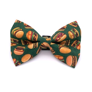 May include: A green bow tie featuring a pattern of colourful hamburgers. The bow tie is made of fabric and has a black adjustable strap.