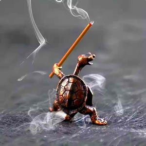 May include: A bronze-colored turtle incense holder with a detailed shell and a stick of incense. The turtle is holding the incense stick, and smoke is rising from the lit end. The holder is on a dark surface.