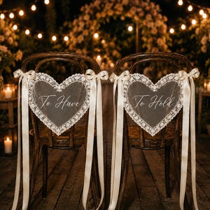 May include: Two dark wood chairs featuring heart-shaped lace decorations attached with cream-coloured satin ribbons. The left heart displays the text To Have, and the right heart displays the text To Hold, both written in a white cursive script font.