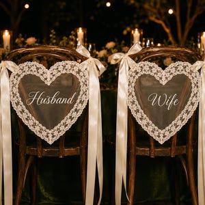 May include: Two heart-shaped chair decorations made of sheer white fabric with intricate lace borders. Each piece features long satin ribbons tied to the chair backs. The left decoration has the word Husband and the right has the word Wife in script.
