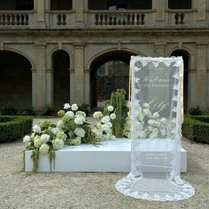 Może przedstawiać: Pionowy, prostokątny znak powitalny na ślub wykonany z przezroczystej białej tkaniny z misternymi koronkowymi brzegami. Tekst brzmi Welcome to the wedding of Lily and David, Tokyo, Japan, 25th April, 2029. Stoi na białej platformie z kwiatami.