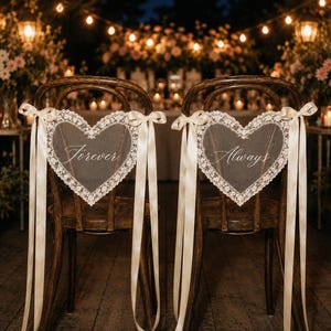 May include: Two dark wood chairs featuring heart-shaped signs with white lace trim. The left sign displays the word Forever in cursive script, and the right sign displays the word Always. Each sign is attached with long, cream-coloured satin ribbons.