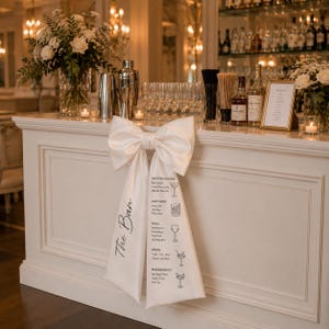 Pode incluir: Um bar branco com um grande laço e um menu. O laço está preso à frente do bar e tem a inscrição "The Bar". O menu lista cocktails, cervejas, vinhos, bebidas espirituosas e refrescos.