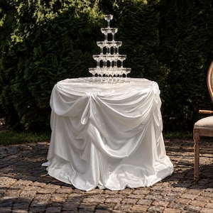 May include: A tiered champagne tower made of clear glass coupes arranged in a pyramid structure, resting on a round table draped in a white satin tablecloth with gathered folds that extend down to the ground, creating a formal display.