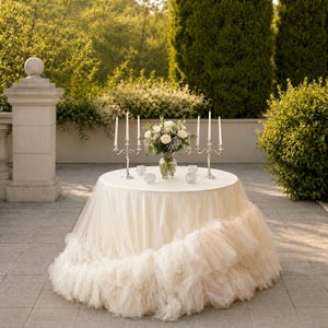 Puede incluir: Una mesa redonda cubierta con una falda de tul de varias capas en color crema con bordes con volantes. La mesa tiene un mantel blanco, un jarrón de cristal central con rosas blancas y vegetación, dos candelabros plateados y pequeños portavelas.