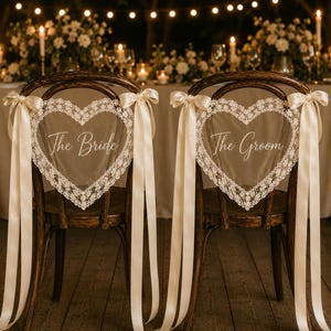 May include: Two dark wood chairs featuring heart-shaped backrests adorned with white lace trim. Each chair displays a sheer heart-shaped sign with white script text reading The Bride and The Groom, accented by long cream-coloured satin ribbons.