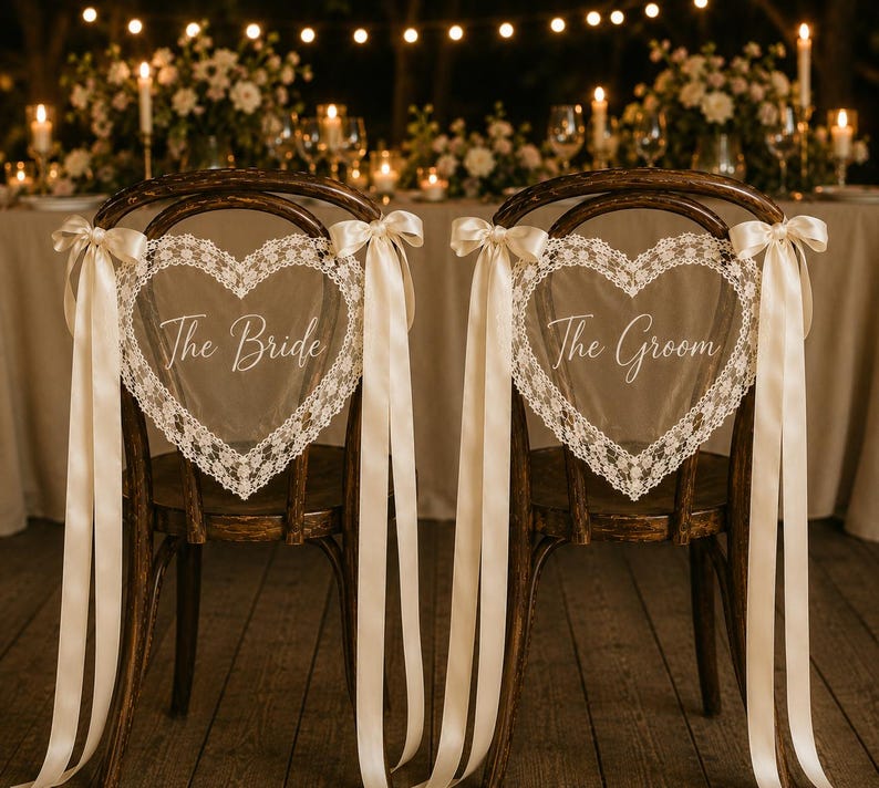 May include: Two dark wood chairs featuring heart-shaped backrests adorned with white lace trim. Each chair displays a sheer heart-shaped sign with white script text reading The Bride and The Groom, accented by long cream-coloured satin ribbons.