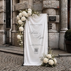 Może przedstawiać: Biały szyld ślubny z tkaniny na czarnej metalowej ramie, ozdobiony białymi kwiatami hortensji. Tekst brzmi: SAVE THE DATE, Samira & Rufus, Sunday, June 25 at 6pm, 123 Anywhere St., Any City, ST 12345, zawiera kod QR z napisem SCAN ME.