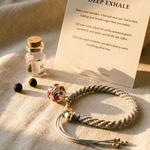 Puede incluir: Una pulsera trenzada con un dije floral plateado y rojo, un pequeño frasco de vidrio con tapón de corcho que contiene cuentas oscuras y una tarjeta color crema con el texto DEEP EXHALE e instrucciones de Tolanda Keepsakes.