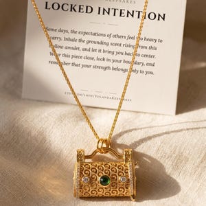Op de afbeelding: Een goudkleurige ketting met een cilindervormige hanger voorzien van filigraanpatronen, een centrale donkergroene cabochon en twee kleine heldere steentjes. De hanger hangt aan een fijne ketting voor een kaart met de tekst LOCKED INTENTION.