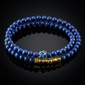 Pode incluir: Duas pulseiras de contas. A pulseira superior é feita de pequenas contas redondas azuis escuras. A pulseira inferior tem as mesmas contas azuis, com contas cúbicas douradas e uma conta decorativa azul e prateada. As pulseiras são exibidas em uma superfície preta.
