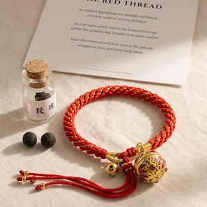 May include: A red and gold braided cord bracelet featuring a spherical gold locket with red enamel details, a small gold bell, and dangling cord ends. Included are two dark incense spheres in a glass vial with a cork stopper and a card.