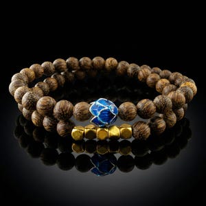 Pode incluir: Duas pulseiras de contas. A pulseira superior apresenta contas de madeira castanha escura. A pulseira inferior tem contas douradas e uma conta azul em forma de flor. As pulseiras são exibidas em uma superfície preta.