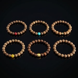 May include: Six wooden bead bracelets with accent beads in red, turquoise, yellow, and gold. The bracelets are made of round, light brown wooden beads. They are displayed on a black surface.