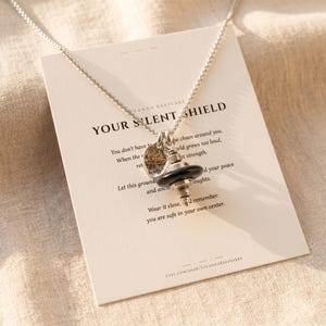 May include: A silver necklace with a pendant featuring a black and silver charm and a small gold-colored charm with a lotus flower design. The necklace rests on a card with the text "YOUR SILENT SHIELD".