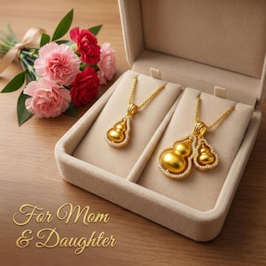May include: Two gold-colored gourd-shaped pendant necklaces with diamond accents are displayed in a beige jewelry box. The box is open, revealing the necklaces. A bouquet of pink and red carnations with green leaves sits to the left. The text "For Mom & Daughter" is at the bottom.