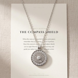 Pode incluir: Um colar prateado com um pingente circular com um design detalhado e uma pedra preciosa central. O pingente pende de uma corrente delicada. O cartão atrás do colar diz "THE COMPASS SHIELD".