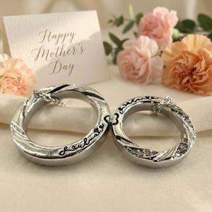 May include: Two silver-toned rings with a twisted design, one with the word "soulmate" and the other with small crystals. A card with "Happy Mother's Day" is in the background, along with pink and peach flowers. The rings are on a white cloth.
