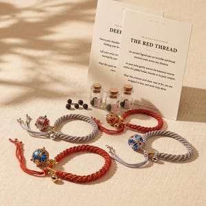 Puede incluir: Cuatro pulseras de cordón trenzado con dijes metálicos, incluyendo dos bandas rojas y dos plateadas. Cada una presenta un relicario esférico con patrones florales esmaltados, pequeñas campanas y cordones colgantes. Se incluyen tres viales de vidrio y dos tarjetas con texto.