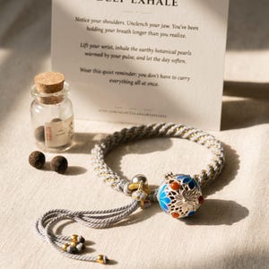 May include: A gray and gold braided cord bracelet featuring a silver-toned filigree locket with blue enamel and orange stone accents. The bracelet includes a tassel end with gold beads, a small glass vial with a cork, and two dark botanical pearls.