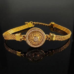 May include: A gold bracelet with a circular centerpiece. The centerpiece is adorned with small, sparkling stones and surrounded by a ring of smaller stones. The bracelet has a chain-like design and is displayed on a reflective surface.