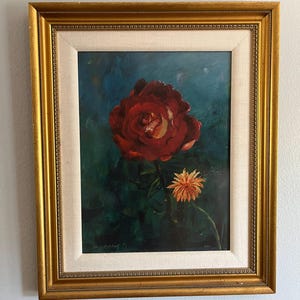 May include: Oil painting of a red rose with a yellow center and an orange flower on a dark green background. The artwork is framed in a gold-colored ornate frame with a white mat.