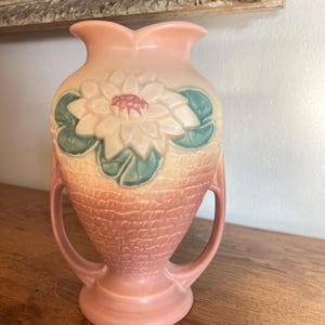 May include: A vintage pink vase with a textured surface and two handles. The vase features a raised floral design with white petals, a red center, and green leaves. The vase is displayed on a wooden surface.