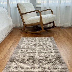 May include: A rectangular, handmade crochet rug with a geometric design in grey and off-white. The rug is displayed on a wooden floor. A rocking chair and a window are visible in the background.