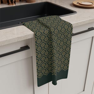 Pode incluir: Uma toalha de cozinha dobrada com um padrão floral repetido em verde-oliva e castanho claro. A toalha está pendurada na borda de uma pia preta, com uma bancada e armários brancos ao fundo.