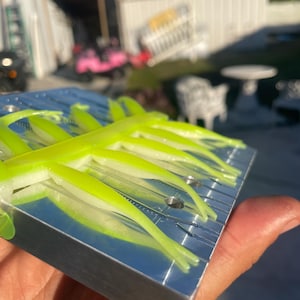 May include: A close-up of a silver metal mold holding several neon green and white soft plastic fishing lures. The lures have a segmented design and are arranged in parallel rows. The mold is held in a person's hand.