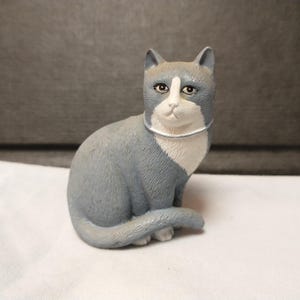 May include: A gray and white ceramic figurine of a cat sitting with its tail curled around its body. The cat has a white collar and black eyes.