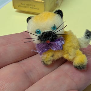 May include: A small, fuzzy, yellow and cream-colored cat figurine with black ears, a black nose, and bright blue eyes. The cat features long black whiskers, a small red mouth, and a purple ribbon tied around its neck. It has a textured, plush appearance.