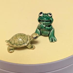 May include: A small ceramic figurine of a green frog sitting upright with its hand near its chin, positioned next to a small ceramic figurine of a light green turtle with a textured shell. Both figures have a glossy finish and painted details.