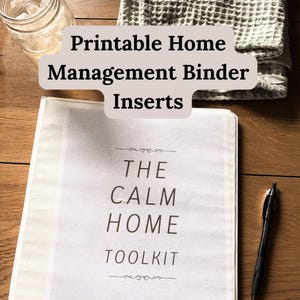 May include: A white binder with a cover page featuring the text THE CALM HOME TOOLKIT in black lettering. The binder is placed on a wooden surface next to a black pen, a glass of water, and a folded grey waffle-textured cloth nearby.