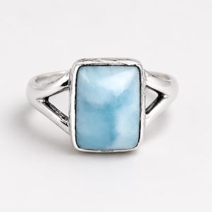 May include: A silver ring featuring a rectangular, light blue, cloudy stone set in a polished metal frame. The band splits into two distinct arms on each side of the central setting, which has a textured, hammered-style edge around the stone.